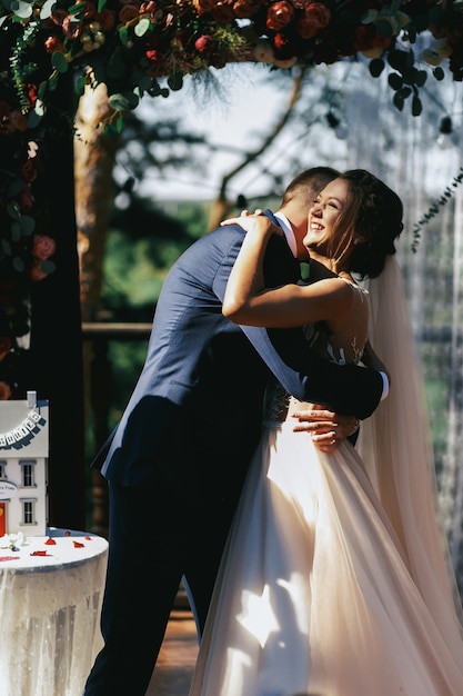 Bruidegom knuffels bruid teder terwijl ze staan ​​onder bruiloft altaar gemaakt van donkere rozen