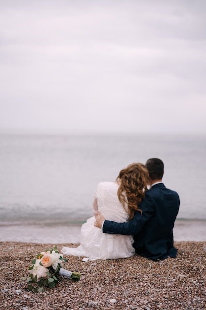 Bruidegom knuffelt bruid in een witte jurk zittend met haar op een kiezelstrand naast boeketten bloemen