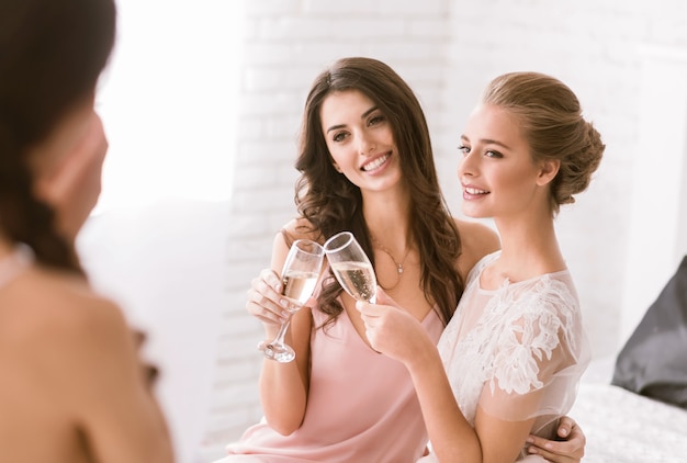 Bruidsmeisjes en bruid met plezier. Blij lachende jonge bruid en bruidsmeisje zitten in de witte kamer terwijl ze champagne drinken en poseren voor de camera