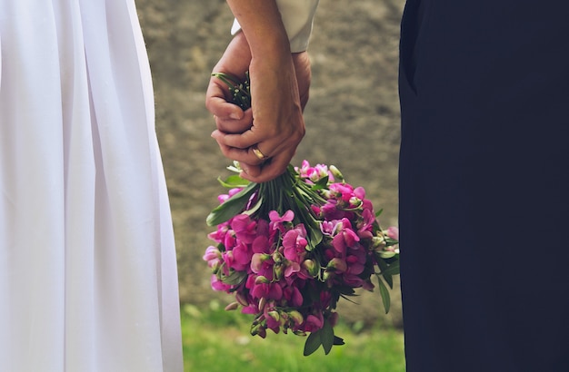 Bruidspaar houdt paars boeket in handen. Romantische zomerfoto van verliefde bruid en bruidegom. Heldergroene gras en steenmuur. Trouwringen en wirwar van armen.