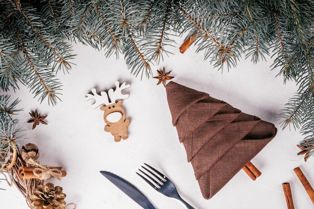 Bruin linnen servet gevouwen in de vorm van kerstboom en mes, vork op wit oppervlak met vuren takken. Bovenaanzicht.