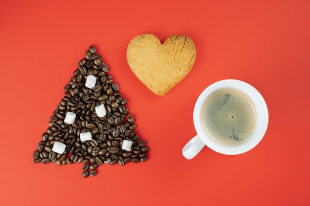 Bruine koffiebonen in de vorm van een kerstboom met een kopje koffie en een hartvormig koekje op een rode achtergrond