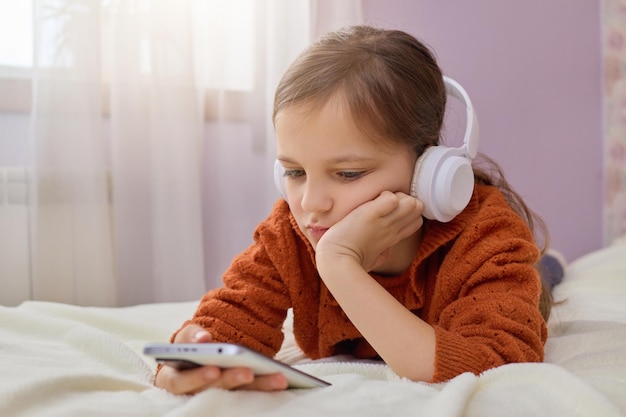 Bruinharig klein meisje in de kinderkamer met koptelefoon en smartphone met mobiele telefoon met headset en thuis video's kijken online