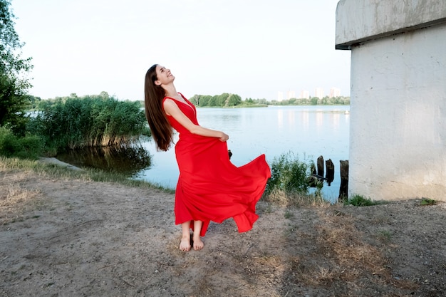 Bruinharige blote voeten meisje met lang haar in rode jurk aan de rivier kust.