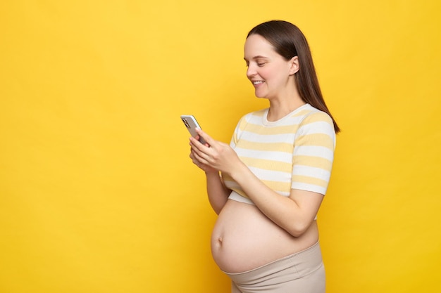 Bruinharige glimlachende blanke zwangere vrouw met blote buik die een casual top draagt geïsoleerd over een gele achtergrond met behulp van een mobiele telefoon internetnieuws lezen online chatten