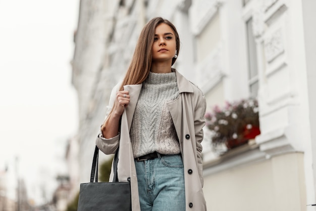 bruinharige vrouw in lente kleding en sneakers met een leren tas poseren langs de straat