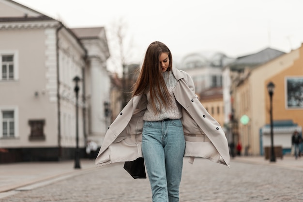 bruinharige vrouw in lente kleding en sneakers met een leren tas poseren langs de straat