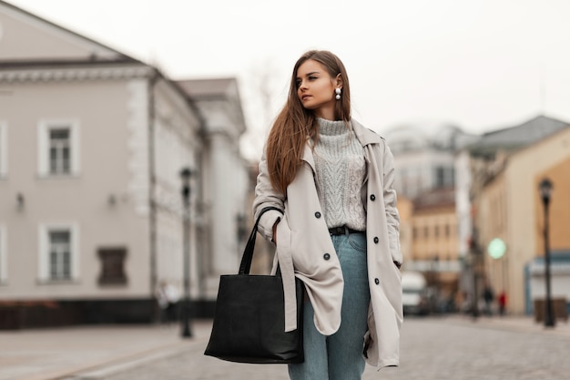 bruinharige vrouw in lente kleding en sneakers met een leren tas poseren langs de straat