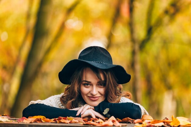 Bruinharige vrouw met hoed glimlacht en baseert haar handen op de tafel met gebladerte in het park