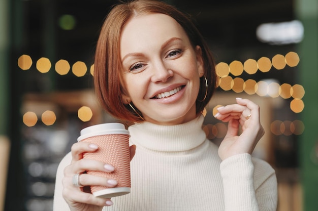 Bruinharige vrouw met make-up brede glimlach gekleed in witte coltrui geniet van warme drank glimlacht positief poseert in coffeeshop heeft make-up drukt geluk uit Drinkconcept