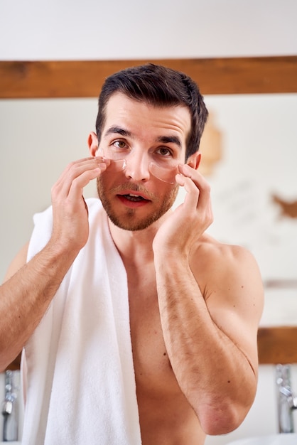 Brunet male applies transparent patches under his eyes while standing in bath opposite mirror in morning