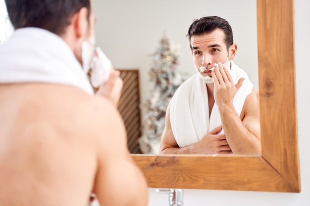 Брюнет с полотенцем на плечах намазывает лицо пеной для бритья, стоя в ванне возле зеркала