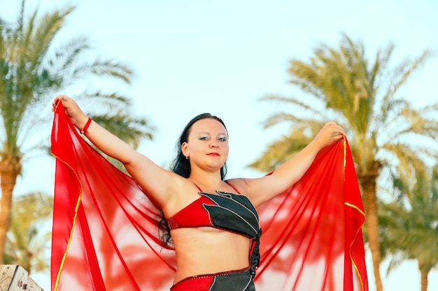 A brunet woman in a red oriental dance costume on a background of palm trees is dancing with a cape.