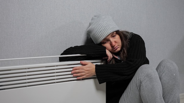 ニット帽とセーターを着たブルネットの女性は、寒さを感じ、ヒーターを抱いて床に座り、暖かく感じます。