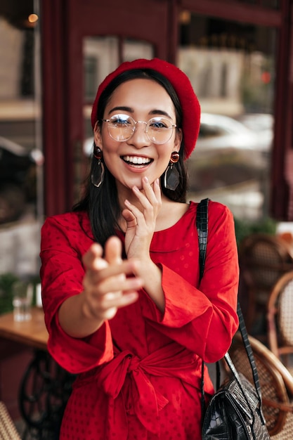 Brunette attractive young woman in eyeglasses red beret and\
bright dress smiles sincerely and pulls hand to camera outside