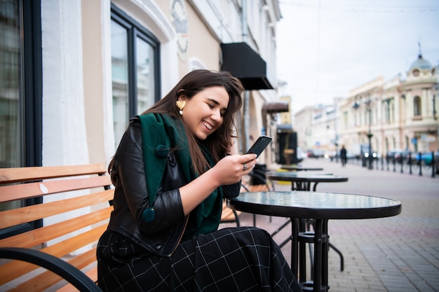 Bella donna caucasica alla moda castana in attrezzatura casuale sulla via europea della città che si siede al messaggio di battitura a macchina del caffè sul suo telefono.