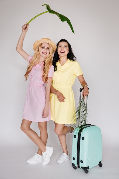 Brunette and blonde with bright dresses on a white wall in a straw hat. Pink and yellow dress. Traveling with a mint suitcase.