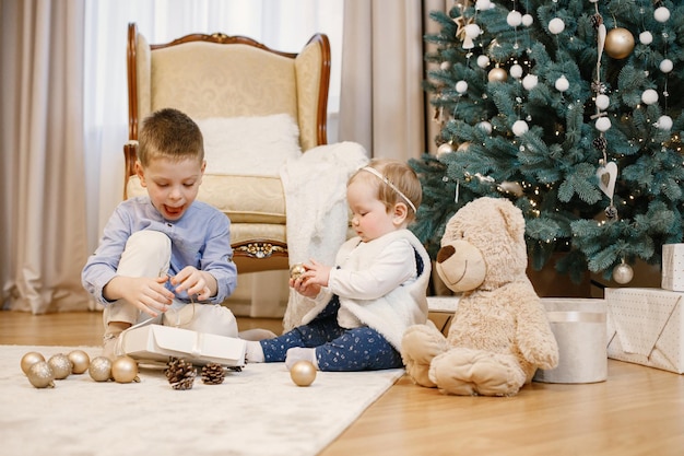Brunette boy and girl sitting near christmas tree at home. Siblings are playing together. Boy and girl wearing blue and beige clothes.