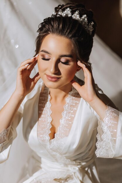 The brunette bride dressed in a satin robe sits on the couch Beautiful hair and makeup Wedding portrait Sincere smile A beautiful bride in a dressing gown is posing