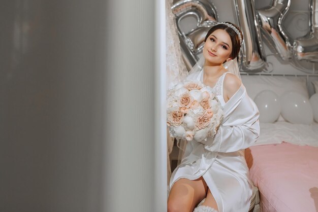 A brunette bride in a white robe and veil poses by the bed with a bouquet of peonies Diadem Wedding concept Morning of the bride Classic hair and makeup