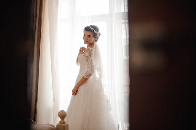 Brunette bruid in een witte jurk staat op een raamscène