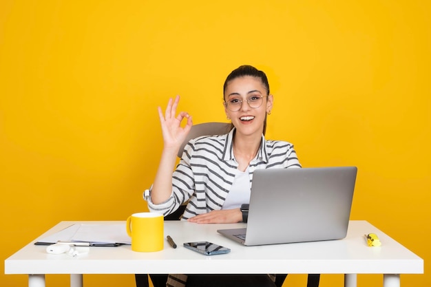 Brunette business woman show ok sign using working on laptop sit office desk over yellow background