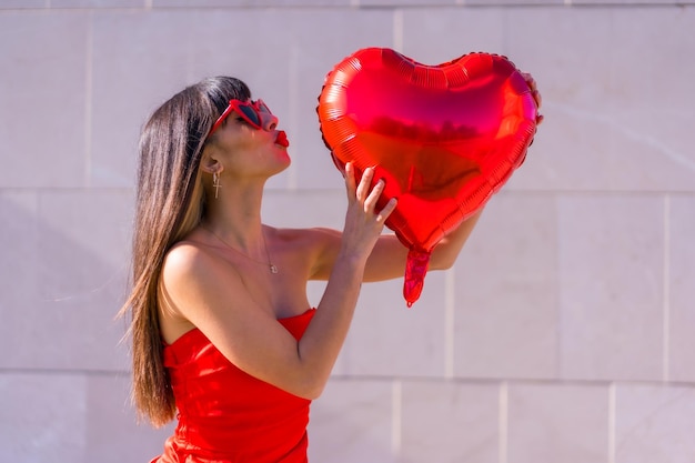 Brunette caucasian girl giving a kiss on Valentines day with a heart balloon sunglasses and red dress