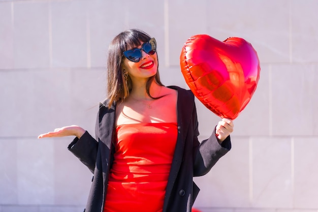 Brunette caucasian girl with a heart balloon on Valentines day sunglasses and red dress Lifestyle of people in love on a gray background smiling