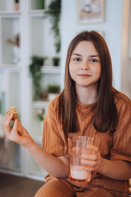 Brunette caucasian teenage girl at home in light brown pajamas holding cookie and glass of milk