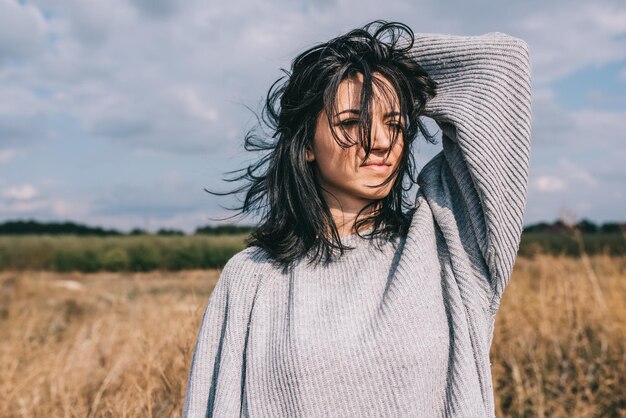 Brunette Caucasian young woman with windy blowing hair outside against meadow and sky Portrait of dream female looking away with hand on head Lifestyle fashion concept Cover idea mood