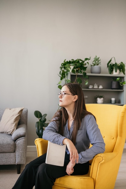 a brunette consultant woman with glasses is sitting in a yellow armchair with a book and looking away