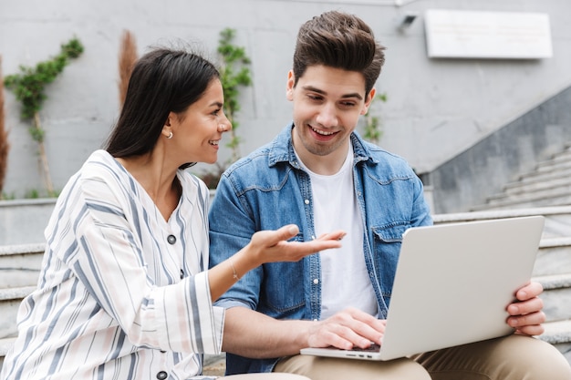 brunette couple man and woman in casual clothes talking and using laptop while sitting on bench near stairs outdoors