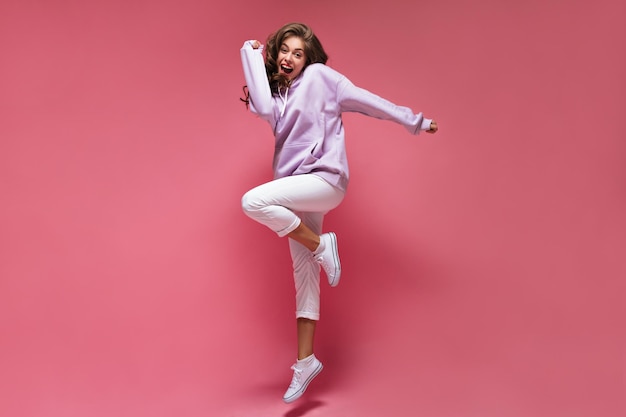 Brunette curly woman in white pants and purple hoodie jumps on isolated Happy girl moves on pink background