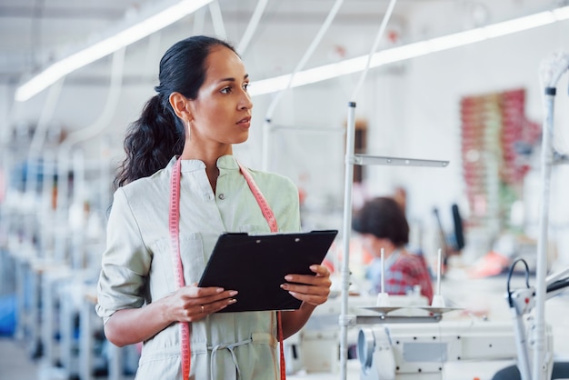 Brunette dressmaker works in the factory and holds notepad in hands.