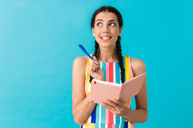 brunette dromende vrouw die omhoog kijkt naar copyspace en schrijft op dagboekboek geïsoleerd op blauwe muur