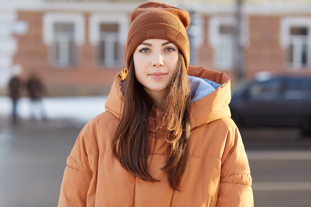 brunette European woman with pleasant appearance, wears hat and brown jacket, looks directly at camera