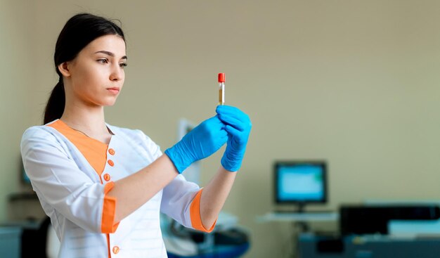 ブルネットの女性医師は、クリニックのぼやけた背景の健康診断の概念の試験管に赤い血を保持しています現代の研究室のクローズアップ