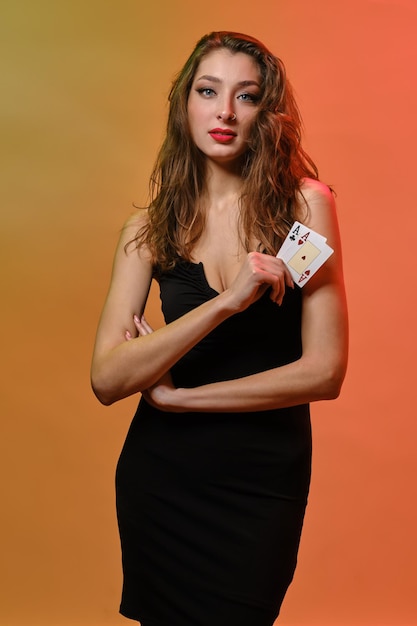 Brunette female with earring in nose in black dress showing two playing cards posing on colorful bac...