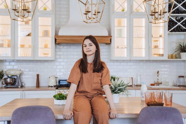 Brunette girl in beige pajamas sitting on the table in the middle of the kitchen looking at camera