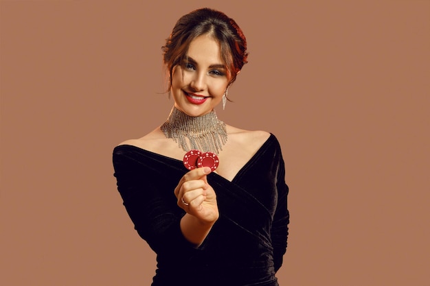 Brunette girl in black dress and shiny jewelry. She smiling, showing two red chips, posing on brown background. Poker, casino. Close-up