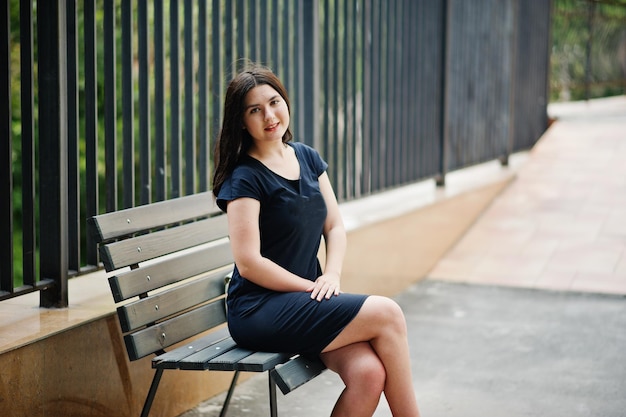 Brunette girl at black dress sitting on bench and posing at street of city