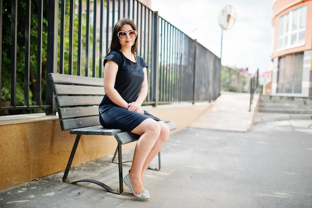 Brunette girl at black dress on sunglasses sitting on bench and posing at street of city