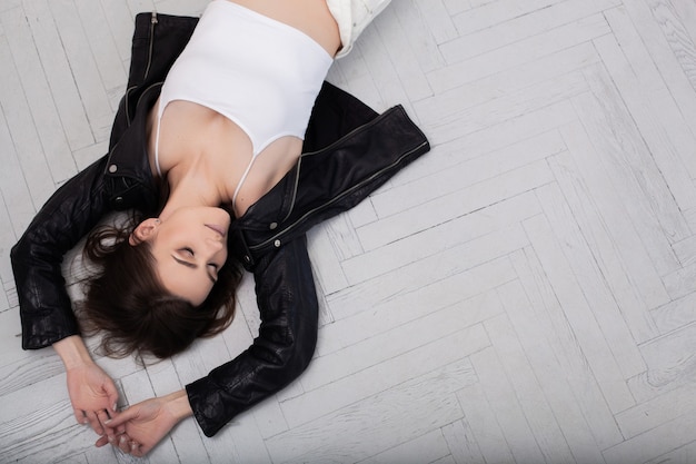 A brunette girl in black leather jacket lying on a white parquet floor background with her hands on the floor over head