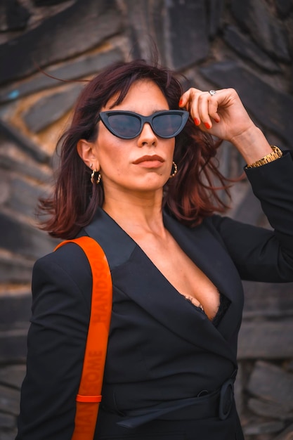 Brunette girl in the city with sunglasses and red bag on a stone wall looking at the camera