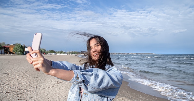 デニムジャケットを着たブルネットの女の子が、海を背景にセルフィーカメラ付き携帯電話で写真を撮ります。旅行と新しい経験の概念。