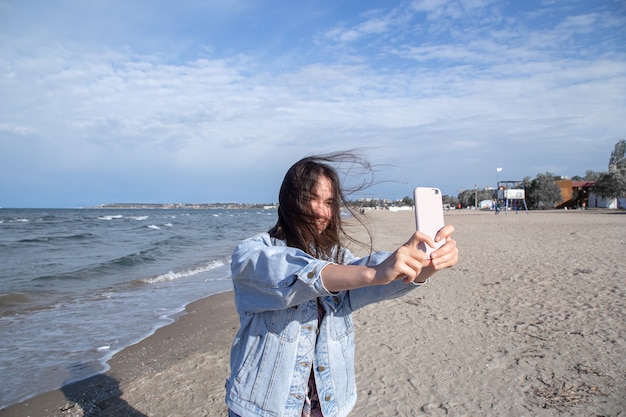 Brunette girl in a denim jacket takes a photo on a selfie camera phone against the wall of the sea. The concept of travel and new experiences.