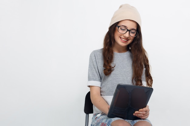 Brunette girl in eye glasses, dressed casual, sitting on chair in studio, holding in her hands a tablet with touch screen