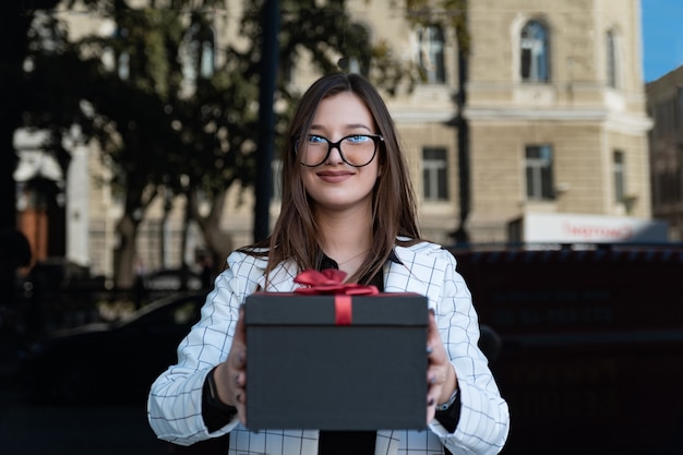 Brunette girl gives you gift. Black box with red bow in female hands. Smiling young woman in glasses holds gift in her hands.