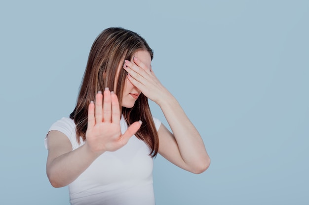 Brunette girl hiding, covered eyes with hand palm isolated over blue wall , portrait. People lifestyle fashion concept. Mock up copy space