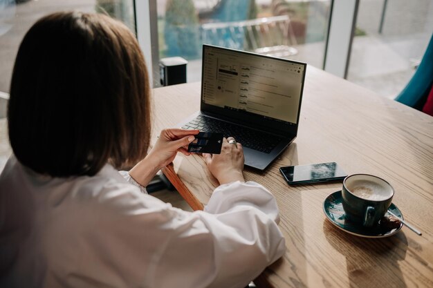 A brunette girl is sitting at a table and shopping online using a plastic card and a laptop